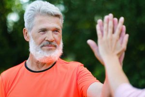 An elderly man in an orange shirt high-fives someone against a blurred nature background.
